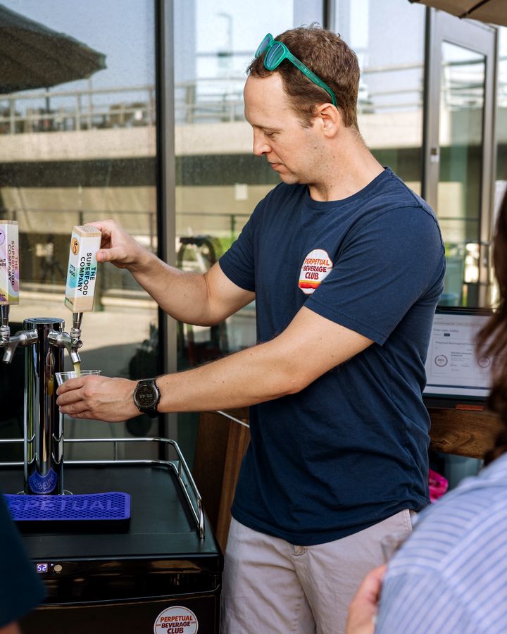 A Perpetual founder pouring The Superfood Company Pink Salt Lemonade kombucha and Passion Fruit Orange Guava probiotic soda from a two-tap Perpetual kegerator at an entrepreneur event in University City